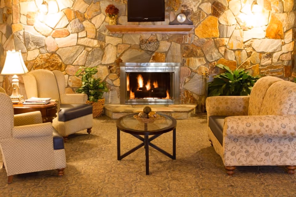 Cozy living room with a stone fireplace in the center, a fire burning inside. There is a wall-mounted TV above the fireplace and a clock on the mantel. Two beige armchairs with patterned upholstery are on the left, next to a wooden side table with a lamp and magazines. On the right, there is a beige floral-patterned sofa. A round glass coffee table with decorative items is in the middle, and green plants are placed near the fireplace.