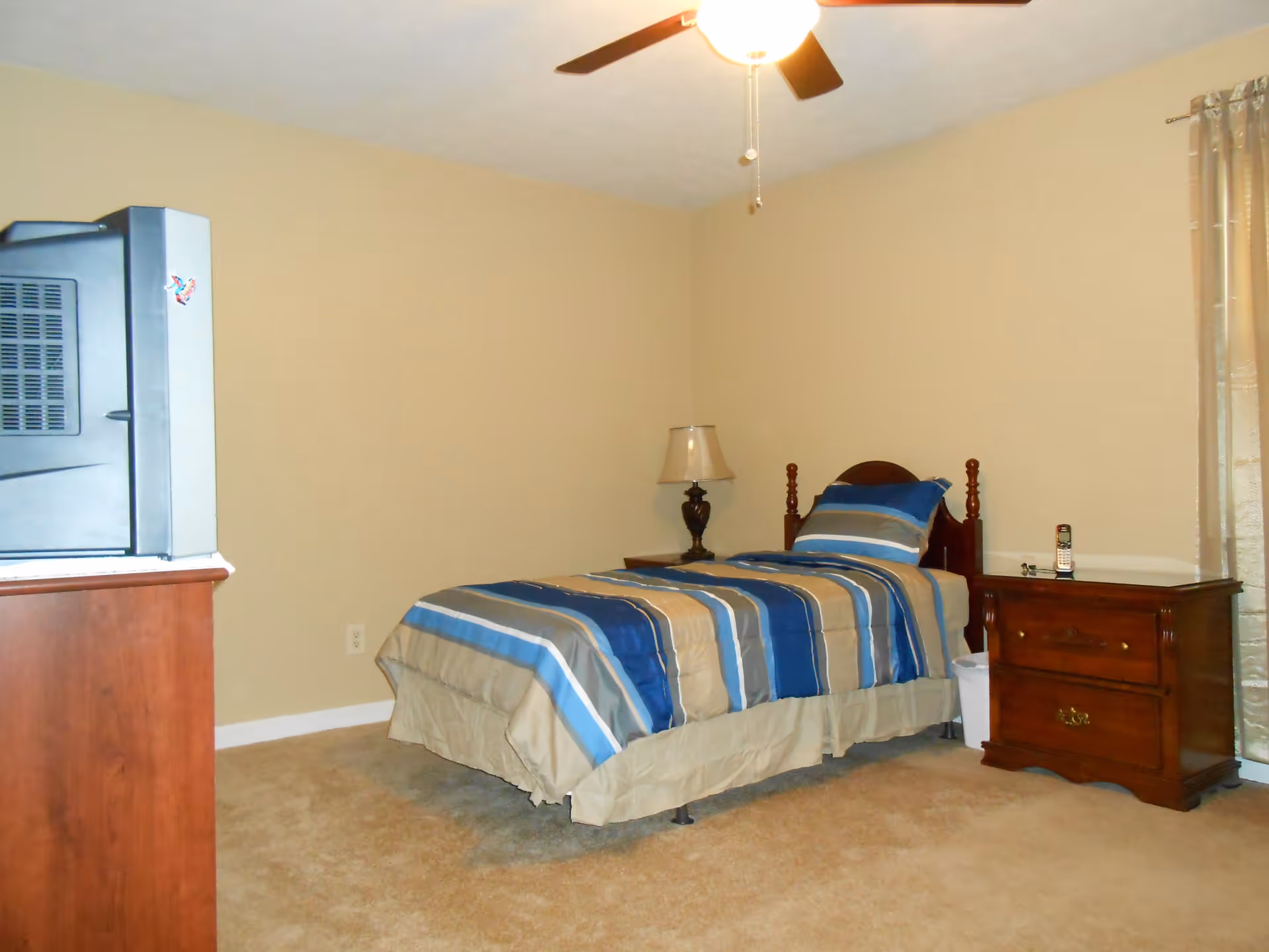 Simple bedroom with a twin bed dressed in blue-striped bedding, a wooden nightstand and dresser, a lamp, and a TV.