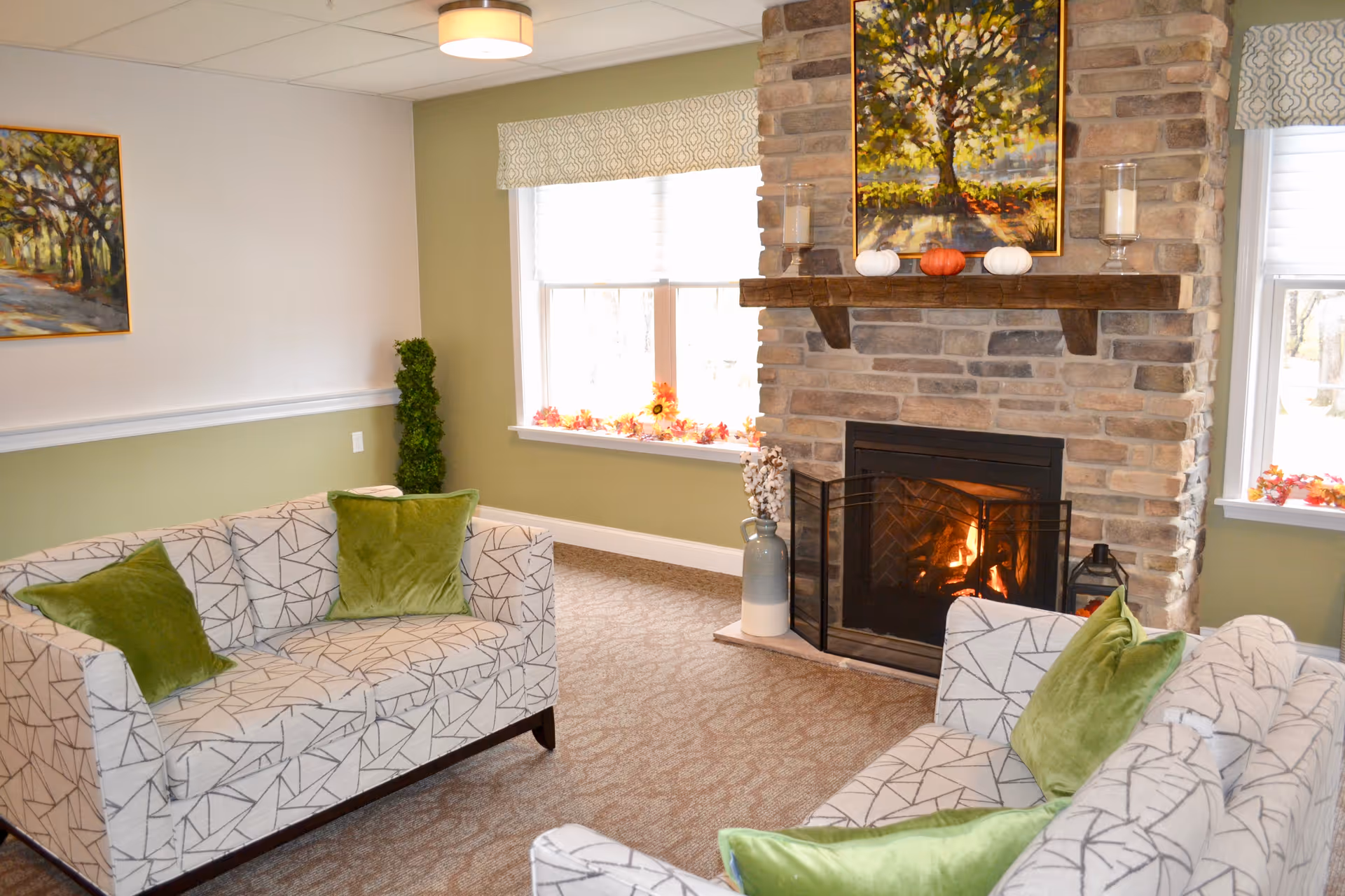A cozy living room with two patterned sofas adorned with green pillows facing a stone fireplace with a fire burning. Above the fireplace is a painting of a tree, and the mantel is decorated with candles and small pumpkins. Two windows with patterned valances let in natural light, and a framed painting hangs on the wall to the left.
