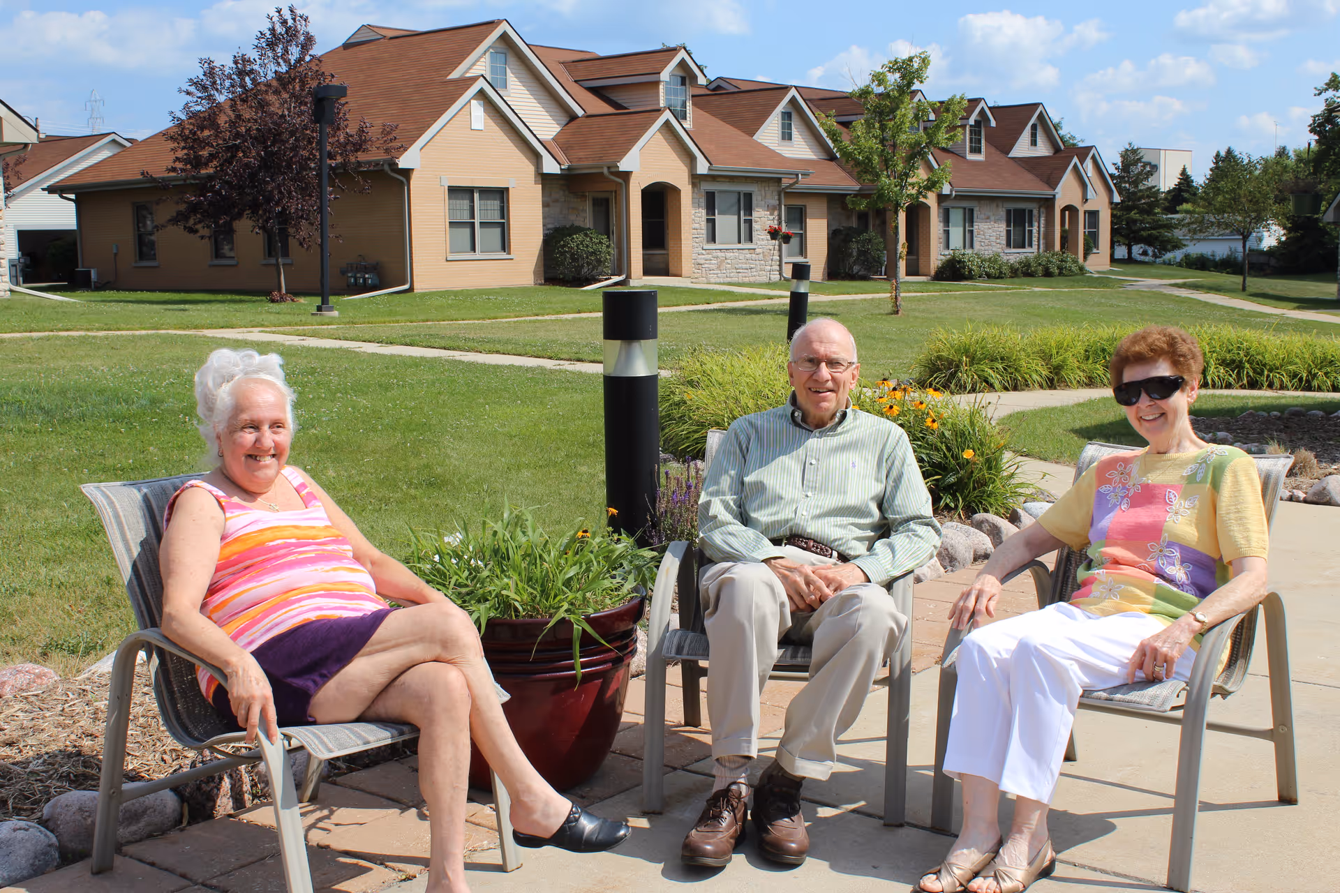 Three elderly people sitting outdoors on chairs in a garden area with green grass, plants, and a paved walkway. Behind them are single-story residential buildings with brown roofs and beige walls under a blue sky with some clouds.