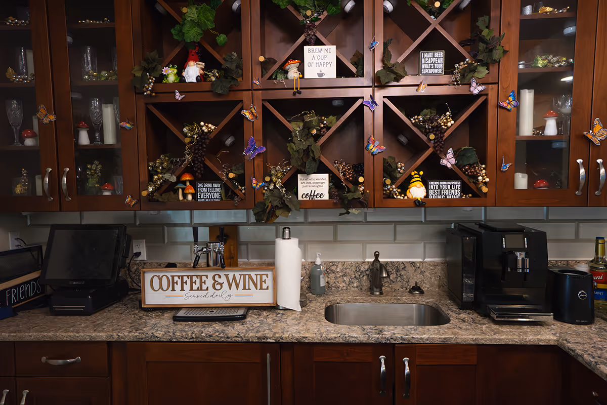 A kitchen countertop with a sink, coffee machine, paper towel holder, and various decorative items. Above the counter are wooden cabinets with glass doors and wine racks filled with artificial grapes and small decorative signs. The signs have phrases related to coffee, wine, and happiness. The backsplash is made of light-colored subway tiles.