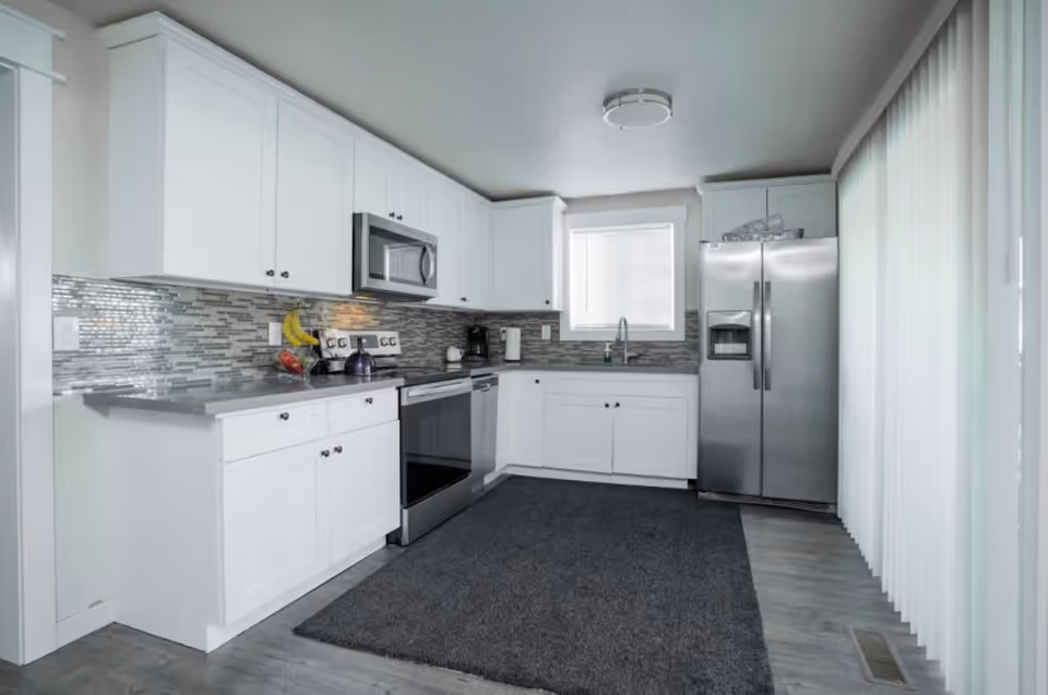 Bright modern kitchen with white cabinets, stainless steel appliances, mosaic tile backsplash, and vertical blinds.