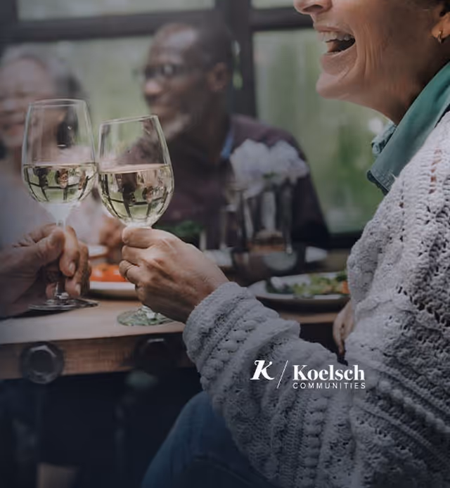 A group of people sitting around a table enjoying drinks and conversation, with two hands holding wine glasses in the foreground and smiling faces in the background.