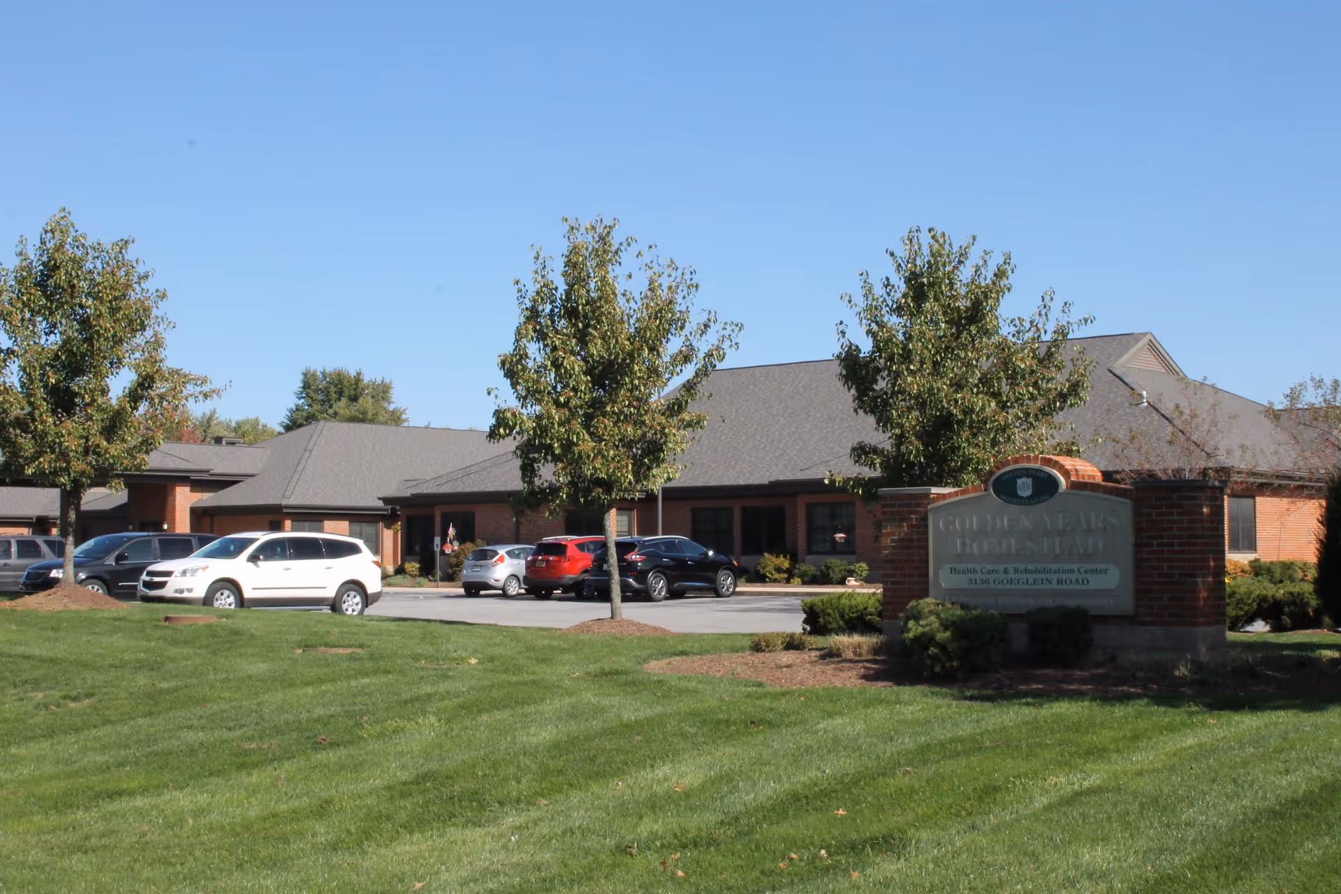 Exterior view of Golden Years Homestead, Inc. showing a single-story brick building with a gray roof, several parked cars, green lawn, and trees under a clear blue sky.