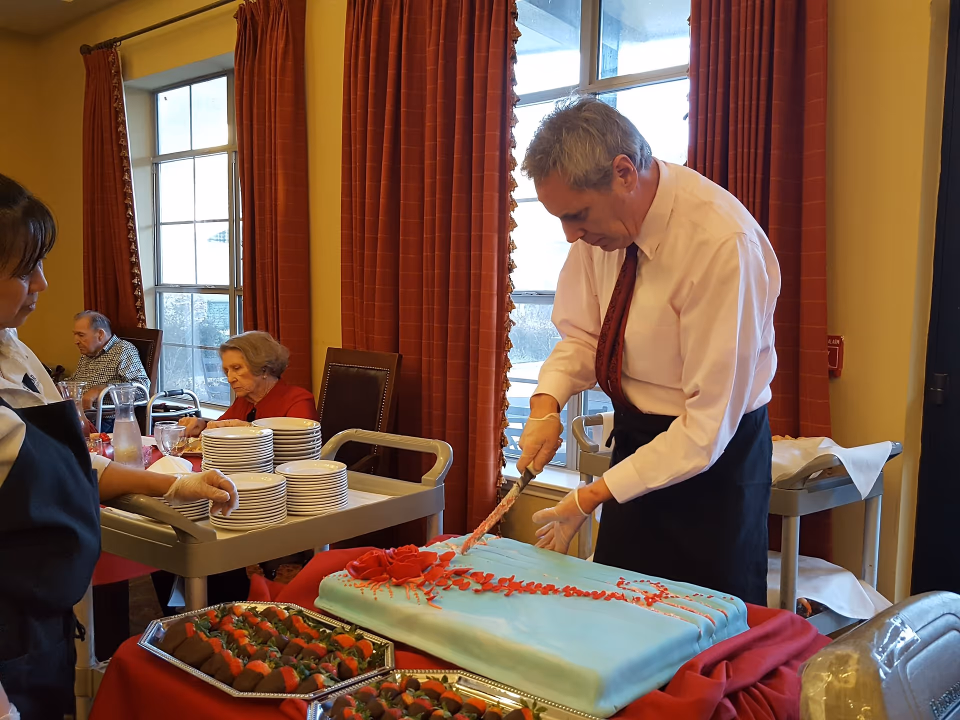 A man in a white shirt and red tie is cutting a large rectangular cake with blue icing and red decorations on a table covered with a red cloth. A woman in a black apron stands nearby holding a tray with stacked plates. In the background, two elderly people are seated near windows with red curtains in a dining area.