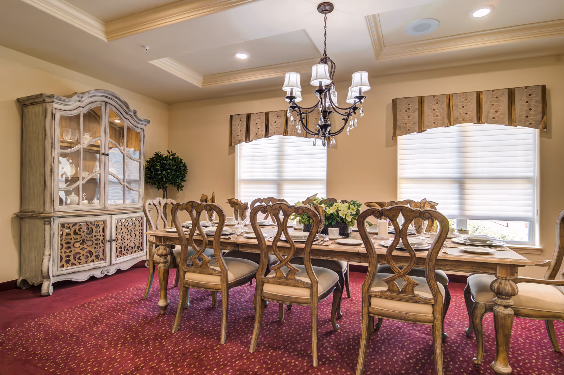 A formal dining room with a wooden dining table set for eight, featuring ornate wooden chairs with cushioned seats. The room has a red patterned carpet, beige walls, two windows with white blinds and decorative valances, a chandelier hanging from a tray ceiling, a large wooden china cabinet displaying glassware, and a potted plant in the corner.