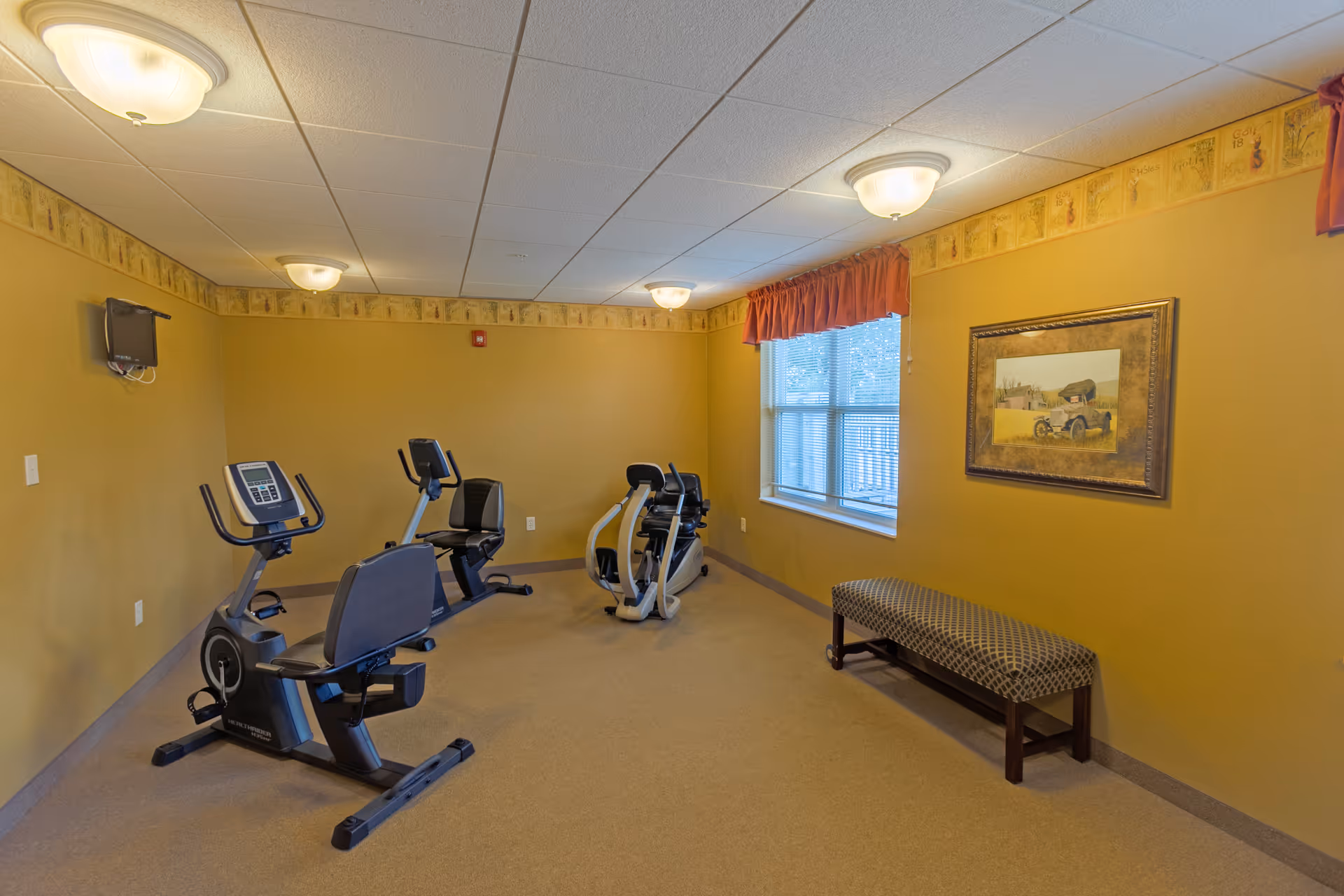 A small exercise room with three stationary exercise bikes, a bench with patterned upholstery, a window with orange valance curtains, a framed painting on the wall, and ceiling lights. The walls are painted yellow with a decorative border near the ceiling.
