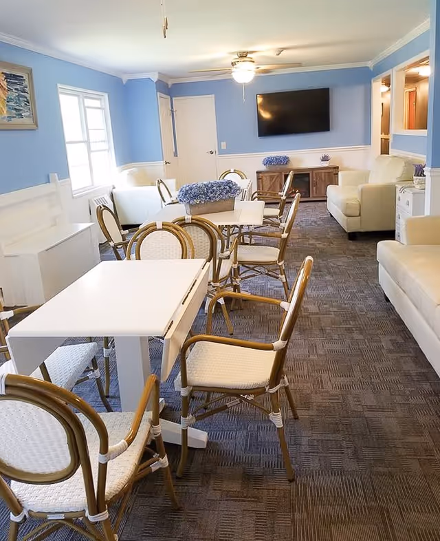 A bright and cozy senior living common area with light blue walls and white trim. The room features several white tables with wicker chairs arranged around them, a ceiling fan with a light, a wall-mounted flat-screen TV, and comfortable white sofas. There are decorative blue flowers on the tables and a wooden cabinet beneath the TV.