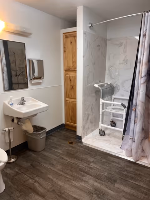 A bathroom with a white sink, a mirror above it, a soap dispenser, a trash can, and a toilet paper holder. There is a walk-in shower with a curtain and a white shower chair with wheels inside. The floor has dark wood-like tiles, and there is a wooden cabinet door in the background.