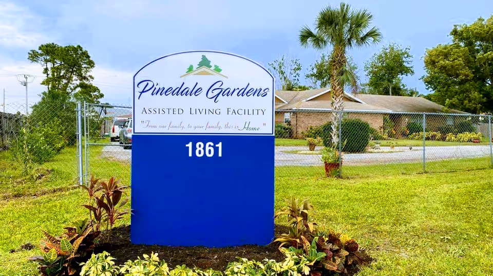 Outdoor view of a sign for Pinedale Gardens Assisted Living Facility with the address number 1861, surrounded by grass, plants, and trees, with a building and a chain-link fence in the background.