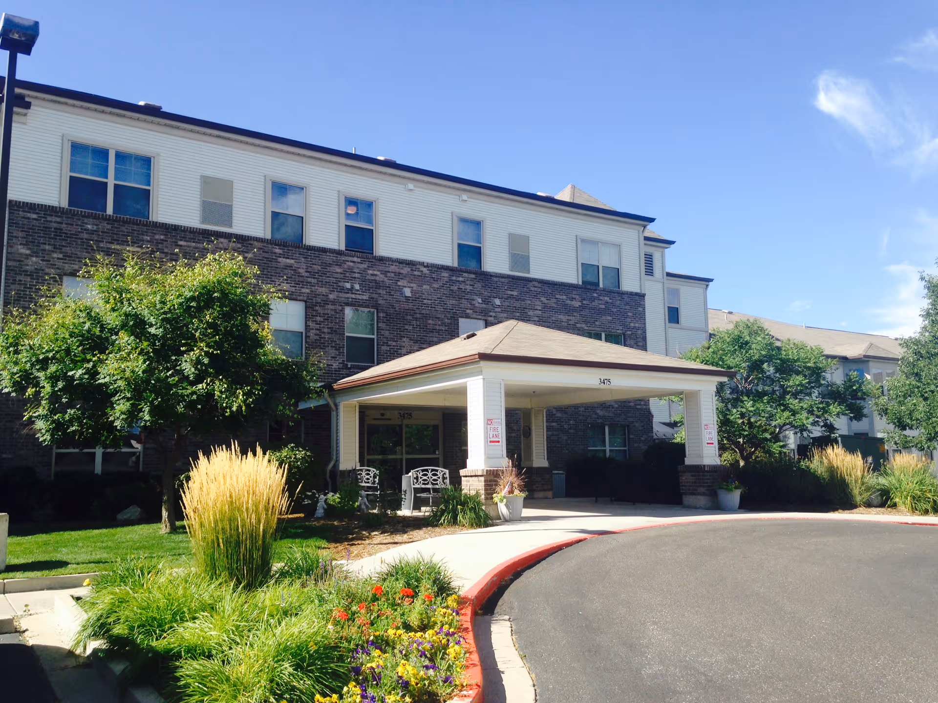 Front exterior view of The Belcaro senior living facility showing a three-story building with a covered entrance, surrounded by green trees, shrubs, and colorful flowers under a clear blue sky.