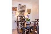 A dining area with a dark wooden table and four matching chairs. The table has decorative items including a potted orchid and a small sculpture. The wall behind the table features four framed artworks and two table lamps with beige lampshades on a sideboard.