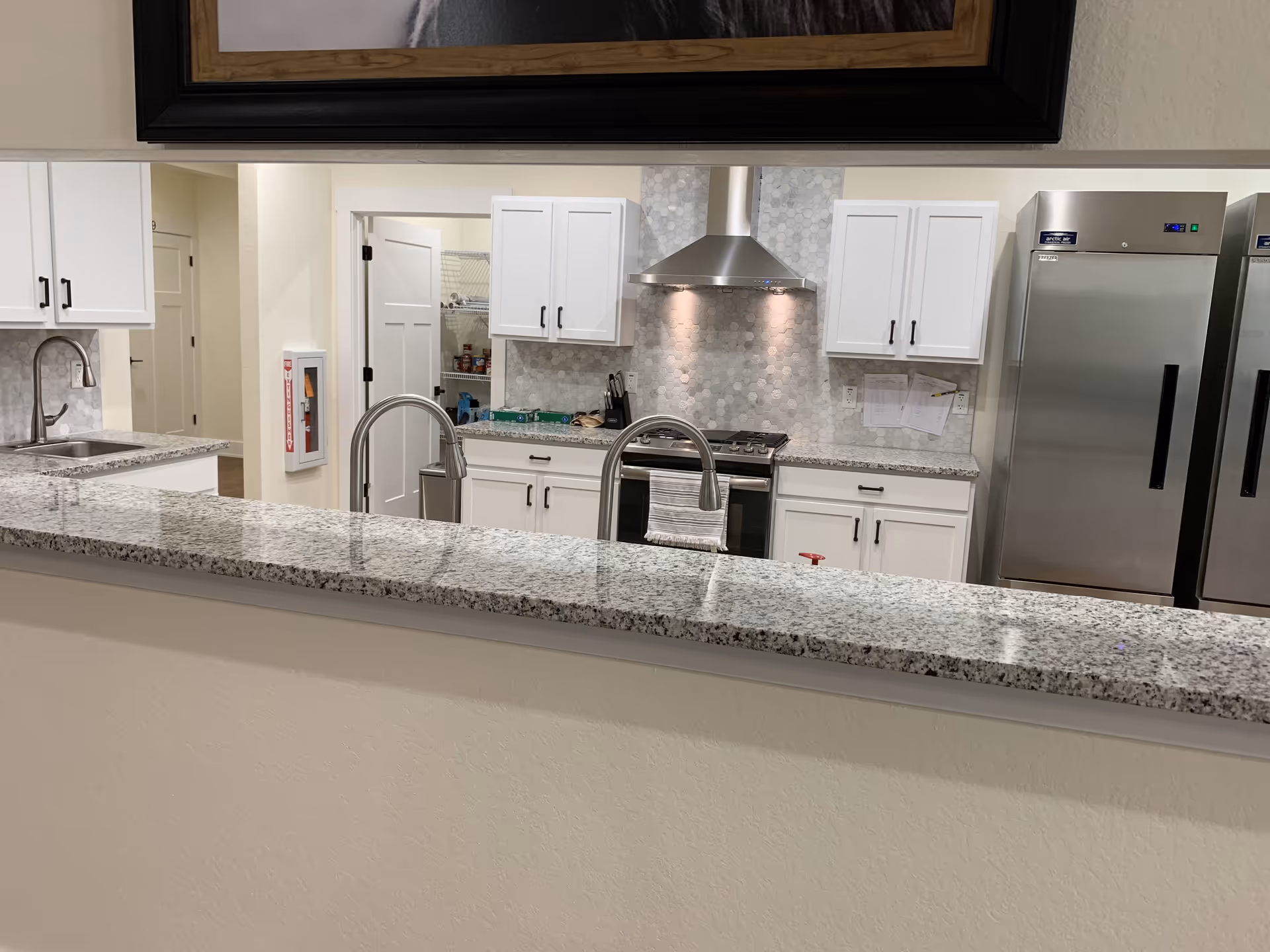 A clean communal kitchen with a granite pass-through counter, stainless steel appliances, white cabinets and twin sinks.