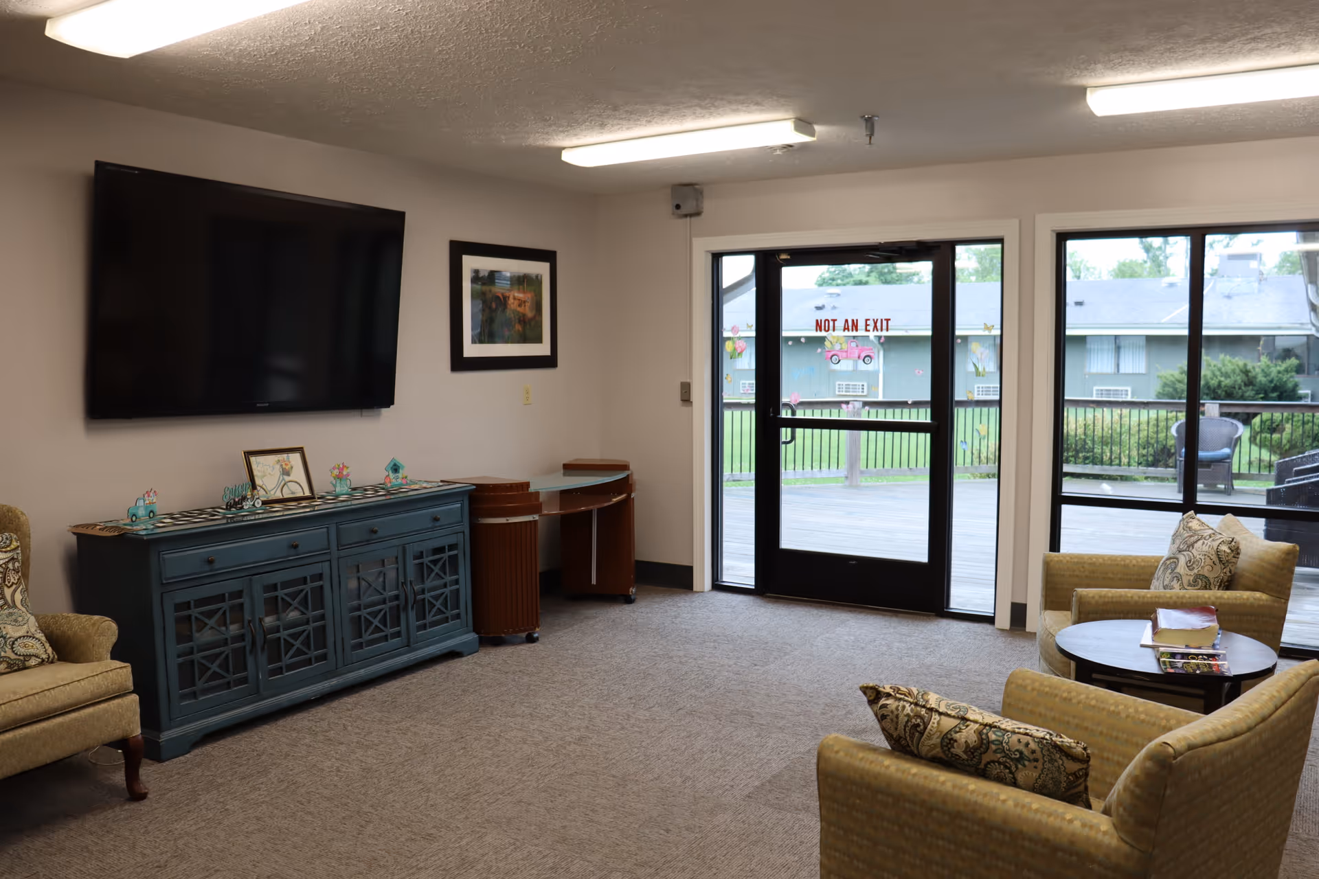 A cozy living room area with a large flat-screen TV mounted on the wall above a blue cabinet. There are two beige armchairs with patterned cushions around a small round table with books on it. A glass door and windows provide a view of an outdoor deck and greenery. The door has a sign that reads 'NOT AN EXIT'.