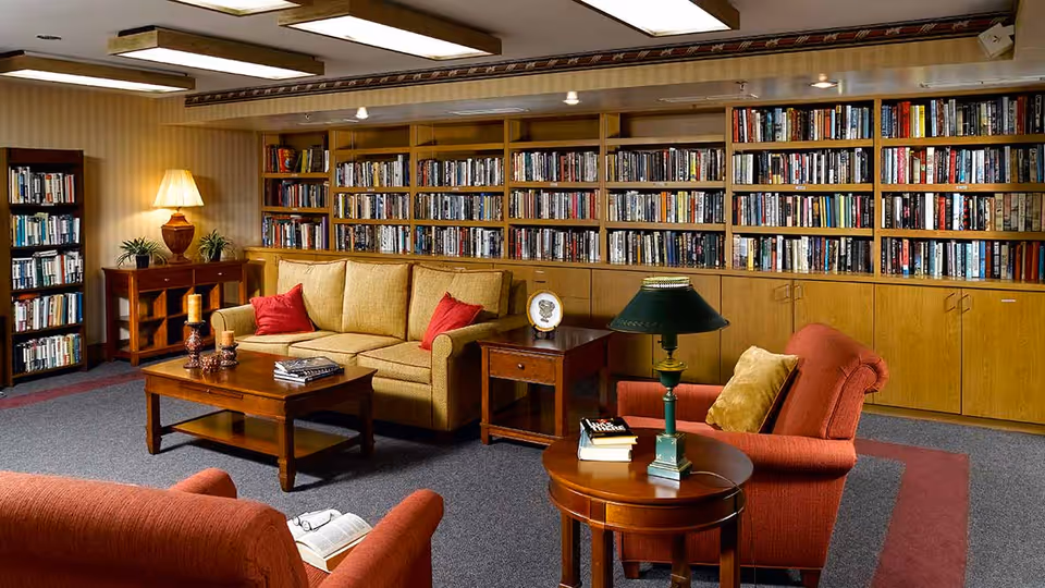 A cozy library room with wooden bookshelves filled with books along the back wall. The room features a beige sofa with red cushions, two red armchairs with a yellow cushion on one, a wooden coffee table with candles and books, a side table with a lamp, and a round wooden table with a green lamp and books. The floor is carpeted with a gray and red pattern, and the walls have a striped wallpaper.