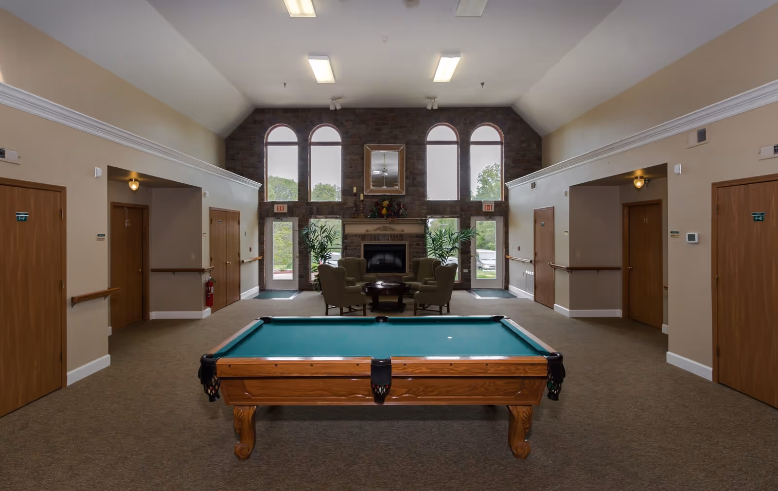 Interior view of a common area in Twin Oaks Estate featuring a pool table in the foreground, a seating area with four armchairs around a coffee table near a stone fireplace, large arched windows letting in natural light, and several wooden doors along the beige walls.
