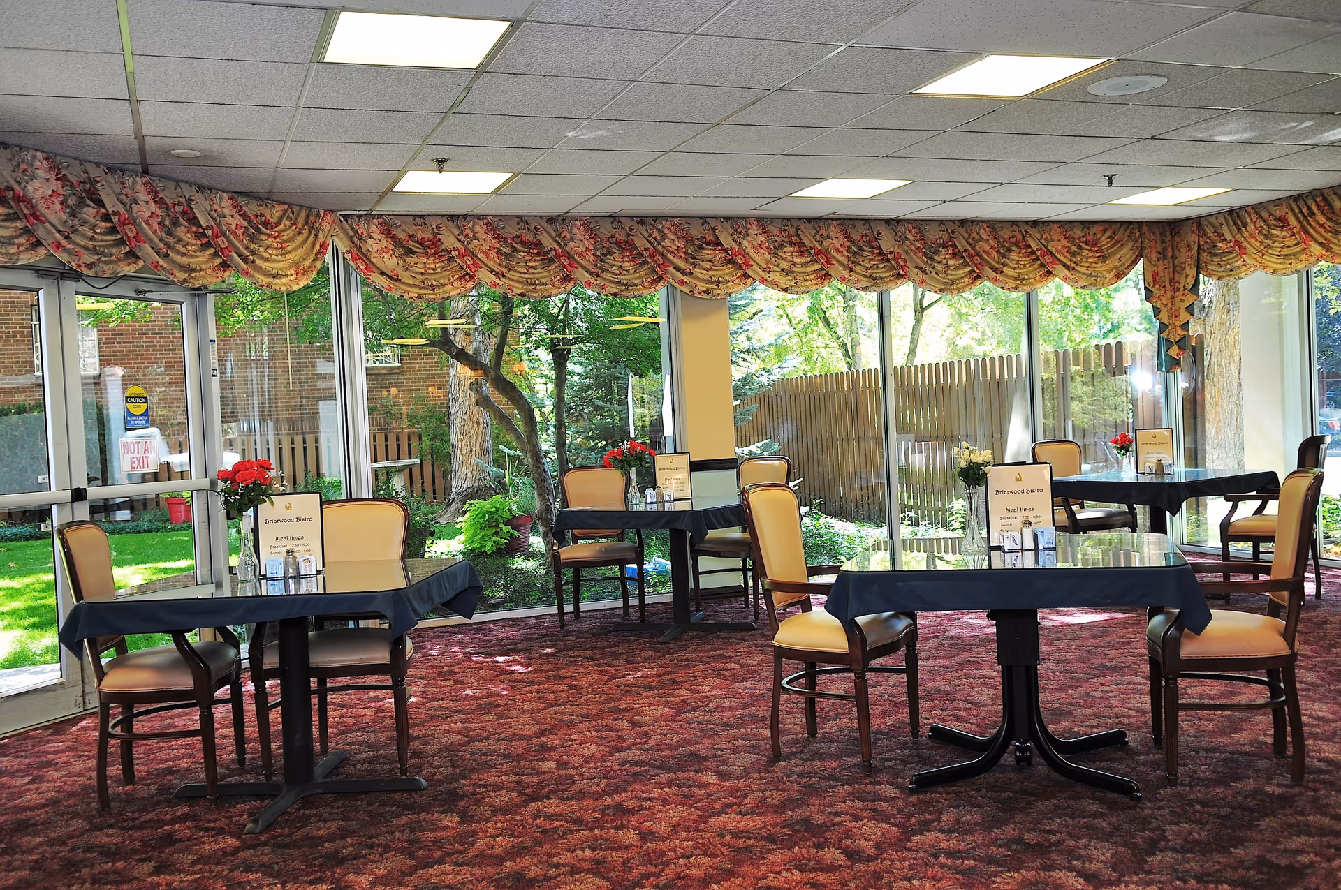 Bright dining room with several tables and chairs, large floor-to-ceiling windows showing a garden, and floral valances.