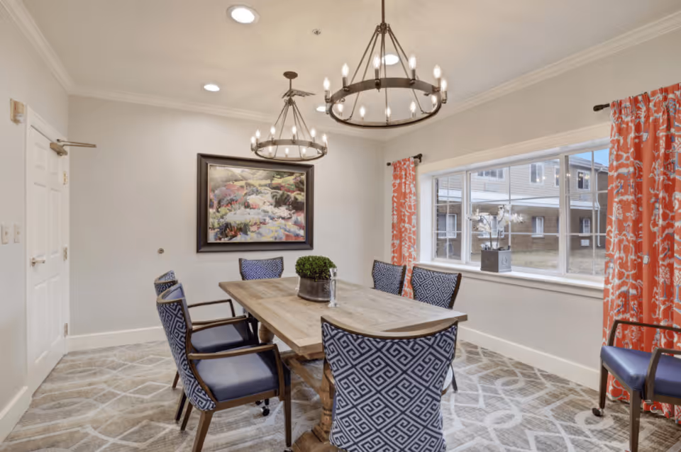 A dining room with a wooden table surrounded by six blue patterned chairs. Two circular chandeliers hang from the ceiling. There is a large window with orange patterned curtains on the right side, and a framed painting on the wall at the far end of the room. The floor is carpeted with a light-colored geometric pattern.