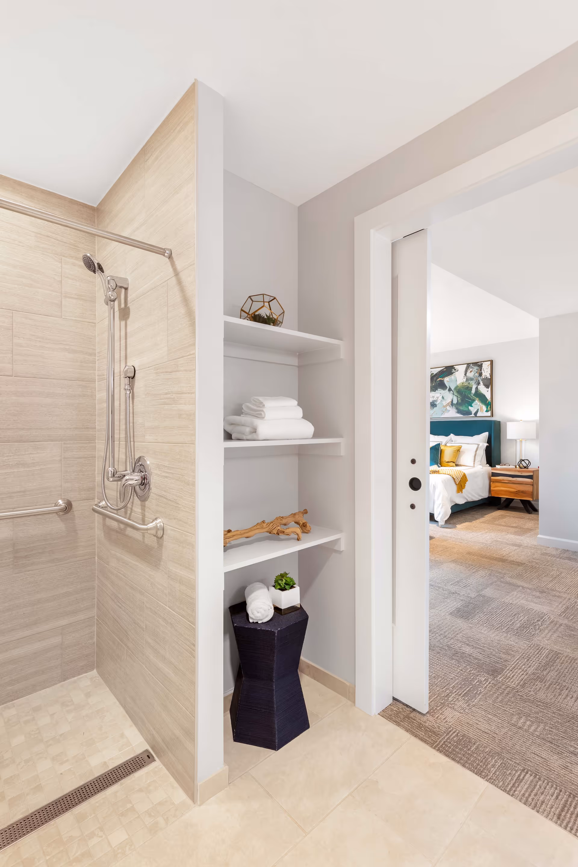 A modern bathroom with a walk-in shower featuring beige tiles and a handheld showerhead. Next to the shower are built-in white shelves holding folded white towels, decorative items, and a small plant. The bathroom opens into a bedroom with a bed, nightstand, lamp, and artwork on the wall.