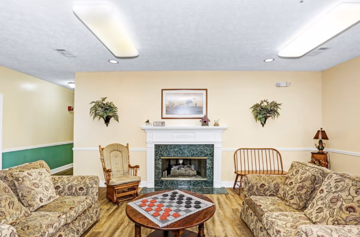 A cozy living room with two patterned sofas facing each other, a wooden rocking chair, and a wooden bench. A round wooden coffee table with a checkers game set is in the center. The room features a white fireplace with a green marble surround, a framed picture above it, and two wall-mounted plants on either side. The walls are painted beige with white trim, and the floor is wood.