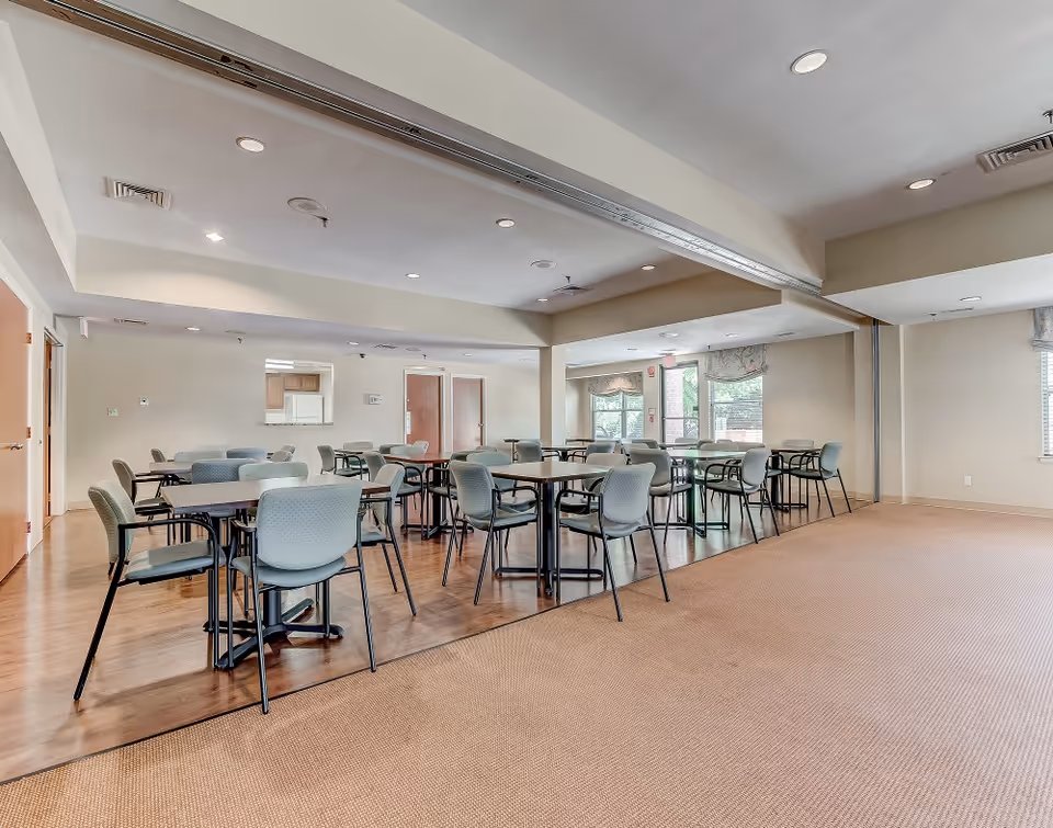 A spacious dining room with multiple tables and chairs arranged neatly. The room has a combination of carpet and wooden flooring, beige walls, and recessed ceiling lights. Large windows allow natural light to enter, and there is a small kitchen pass-through window visible in the background.
