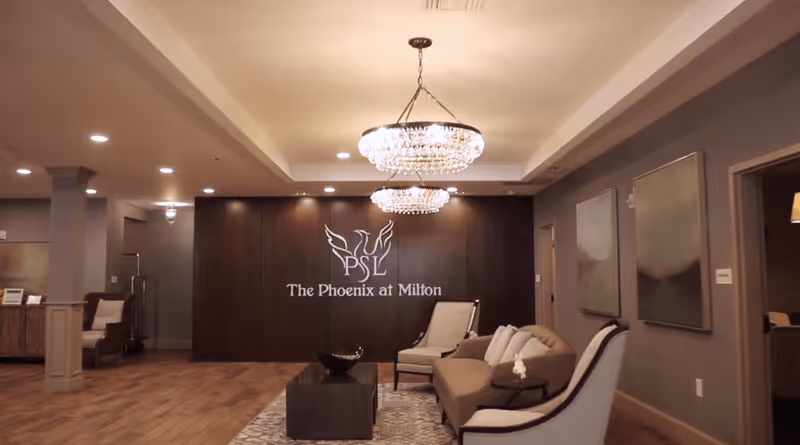A spacious and elegantly decorated living room area with wooden flooring, a beige sofa, two armchairs, a coffee table, and two large chandeliers hanging from the ceiling. The back wall features a dark wood panel with the logo and text 'The Phoenix at Milton'. Two abstract paintings hang on the right wall, and there are doorways leading to other rooms.