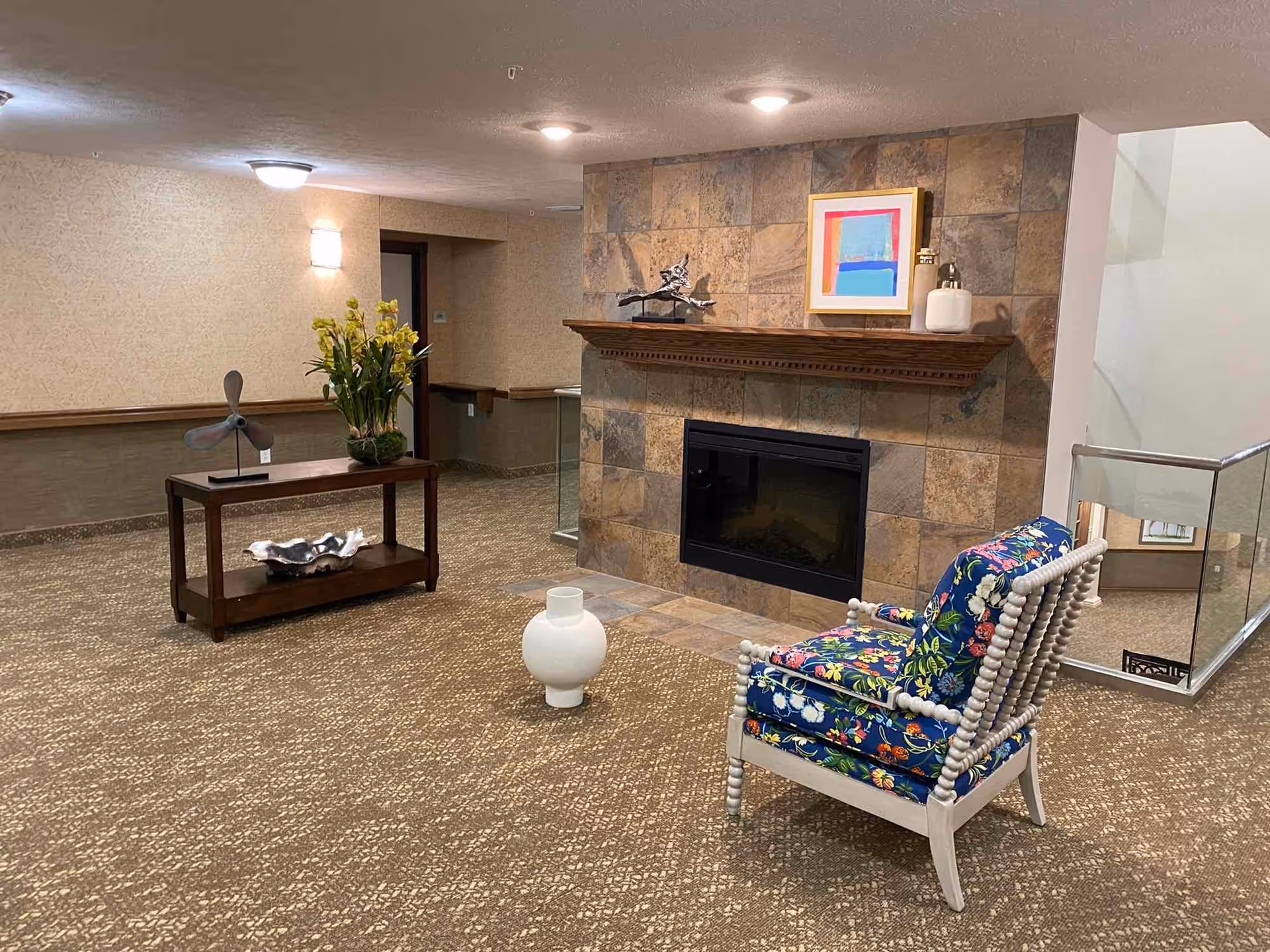 A cozy interior space featuring a stone fireplace with a wooden mantel displaying a colorful framed artwork, a small sculpture, and a white decorative item. In front of the fireplace is a white wooden chair with floral patterned cushions. To the left, there is a wooden console table with a metal fan sculpture and a vase of yellow flowers. The floor is carpeted with a patterned design, and the walls have a textured wallpaper. A glass railing is visible on the right side, indicating a staircase or open area below.