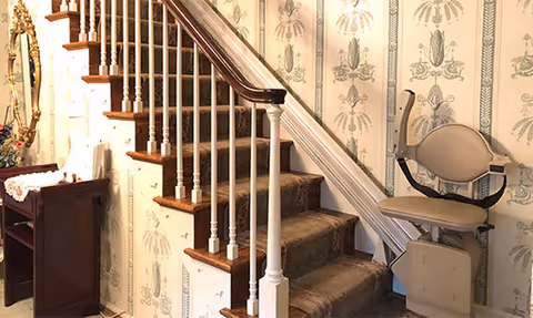 Interior view of a carpeted wooden staircase with a beige stairlift installed, floral wallpaper, and a small side table with a mirror.