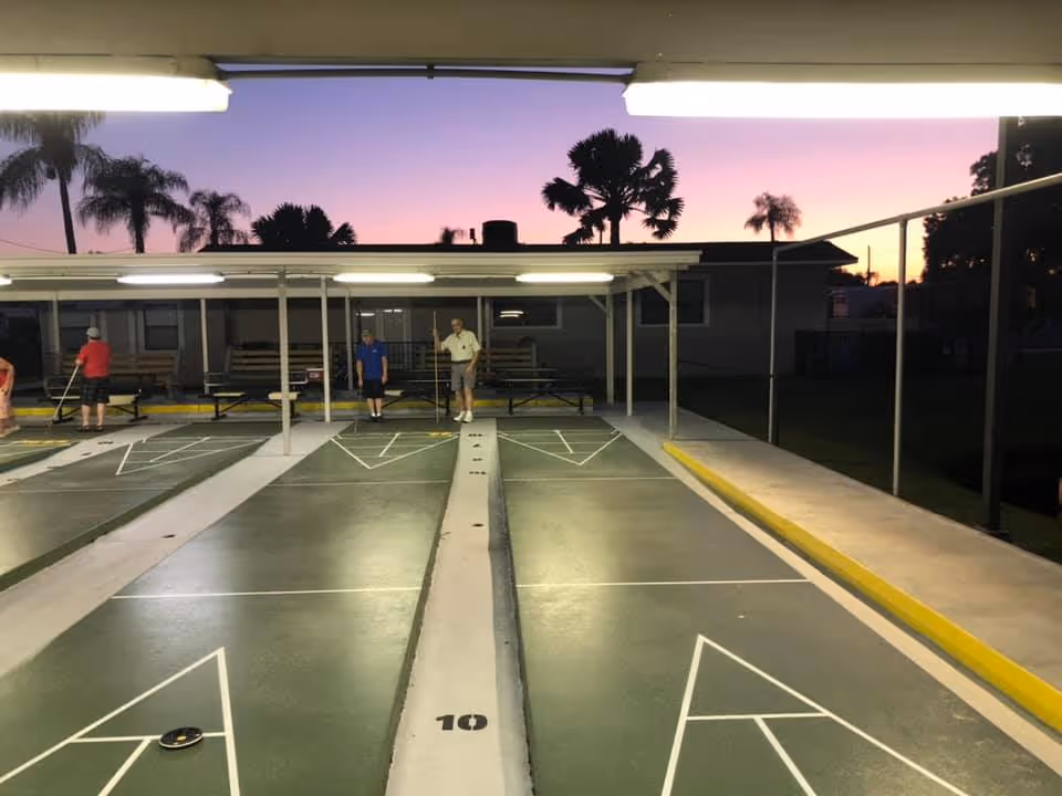 Shuffleboard courts at a retirement community with several players under covered lights at sunset.