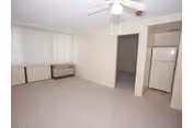 Empty apartment living area with ceiling fan, carpeted floor, vertical blinds, wall air unit, and small kitchenette with refrigerator.