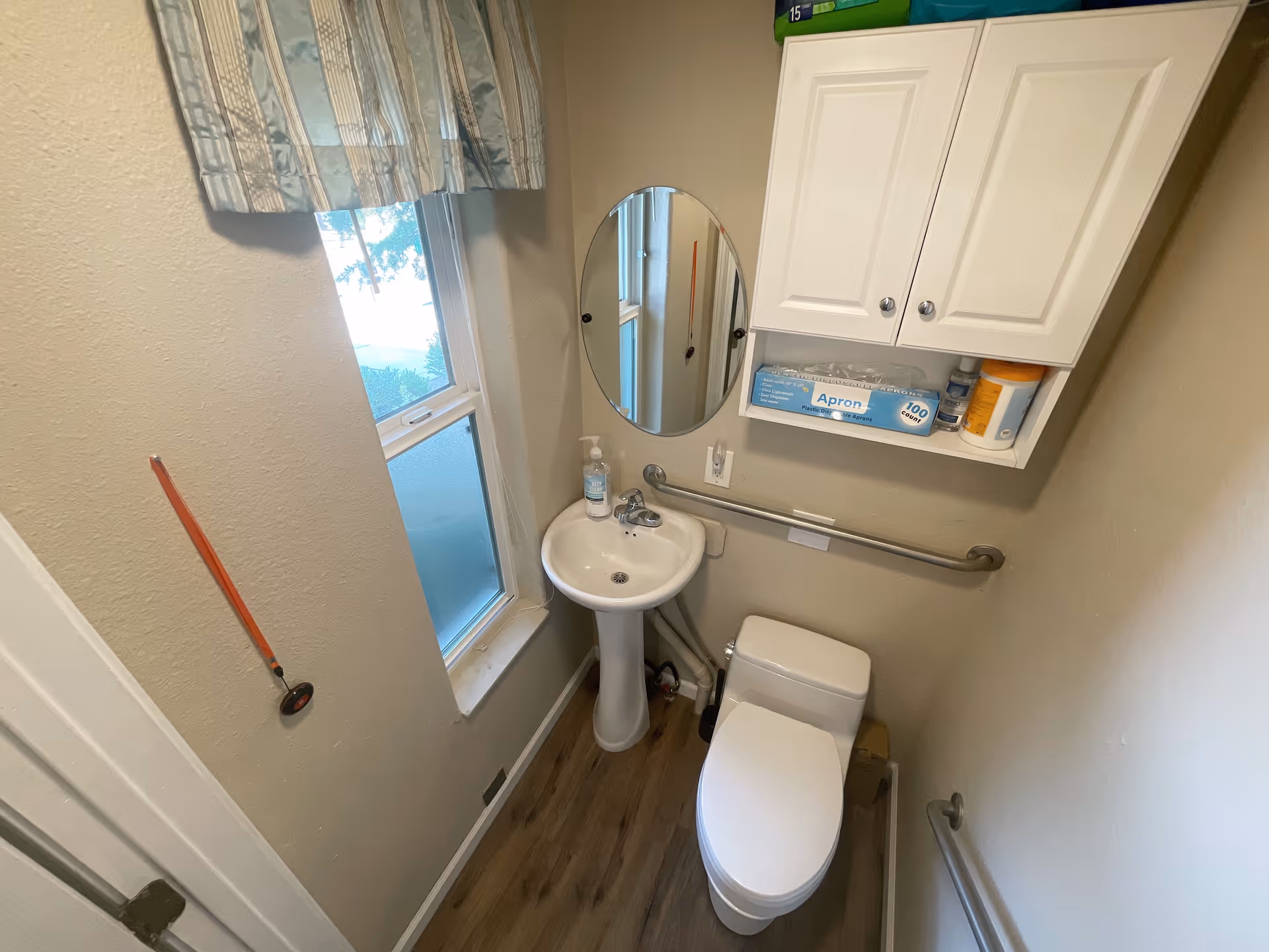 Small bathroom with a white pedestal sink, a toilet, and a round mirror above the sink. There is a window with a curtain on the left wall and a cabinet with medical supplies above the toilet. Grab bars are installed on the walls for support.