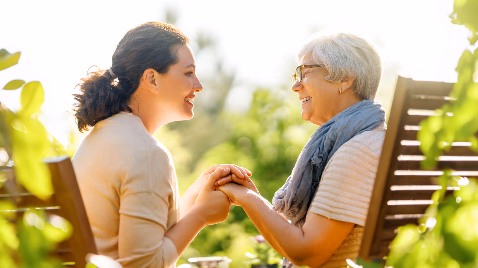 A younger woman and an older woman sitting outdoors on wooden chairs, holding hands and smiling at each other with greenery in the background.