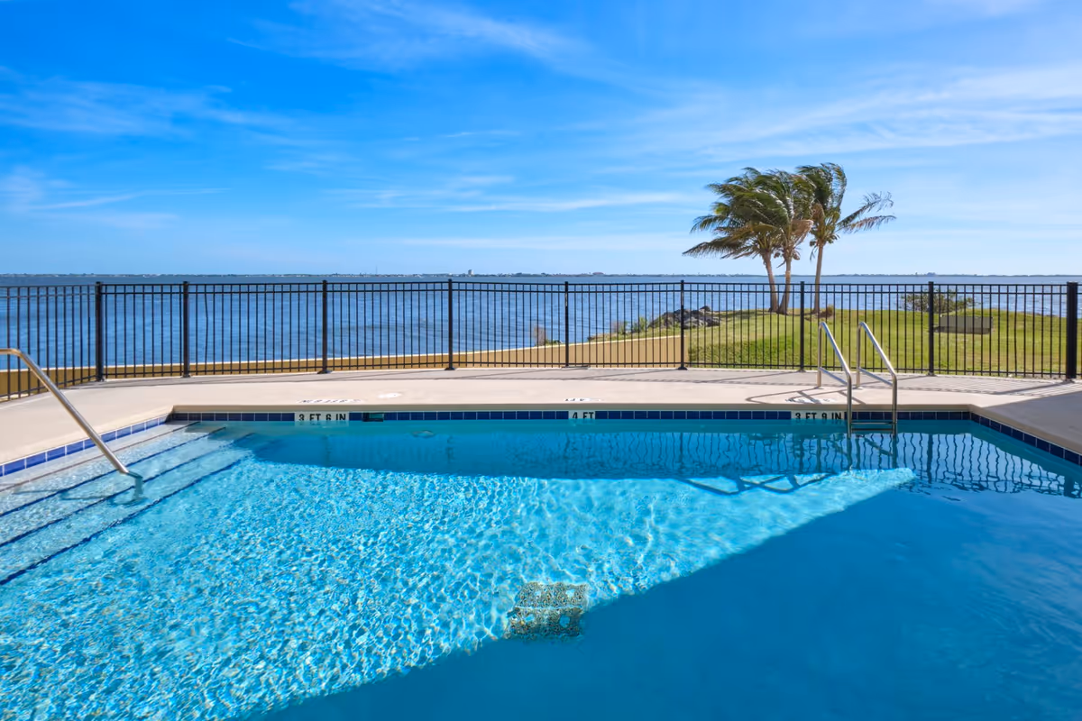 Outdoor swimming pool with clear blue water, metal handrails, and steps leading into the pool. Beyond the pool is a black metal fence, green grass, and two palm trees blowing in the wind. In the background, there is a large body of water under a blue sky with some clouds.