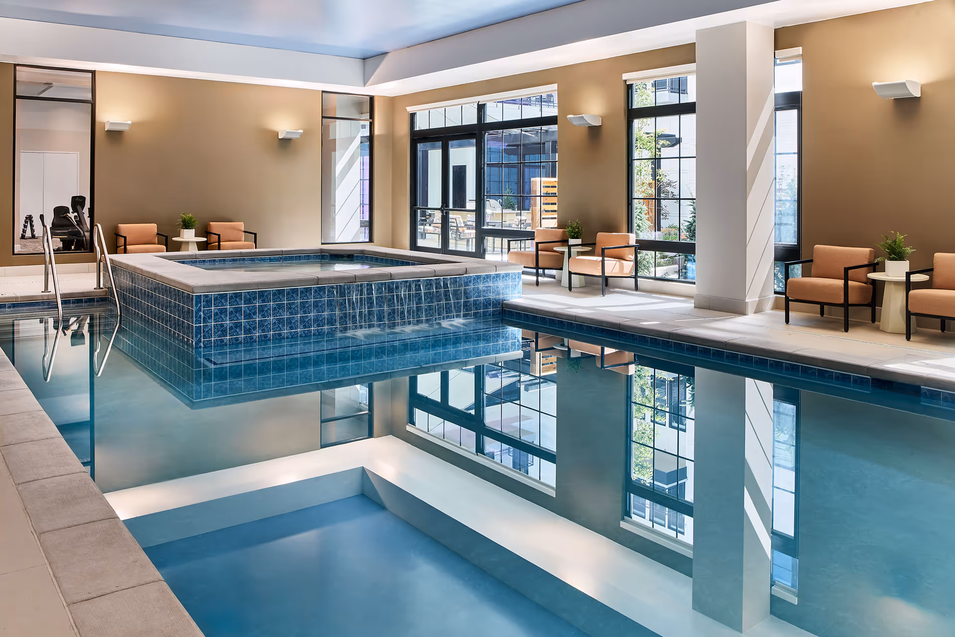 Indoor swimming pool area with a raised hot tub in the center, surrounded by large windows letting in natural light. There are several cushioned chairs and small tables along the walls, and the room has a calm, inviting atmosphere.