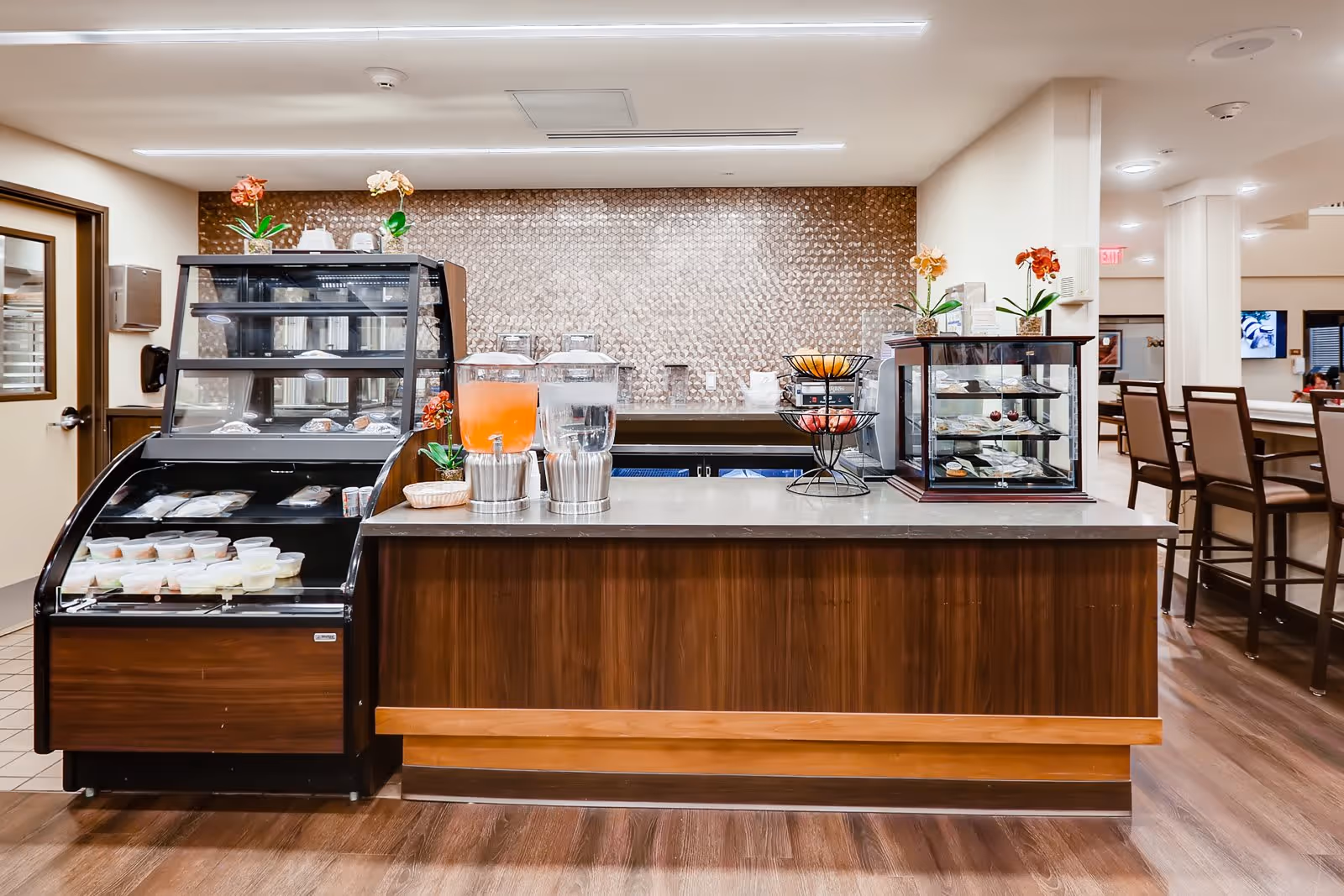 A modern food and beverage station in a senior living facility with a wooden counter, glass display cases containing food items, two beverage dispensers with orange and clear drinks, and a tiered fruit basket. The background features a textured wall and seating area with high chairs.