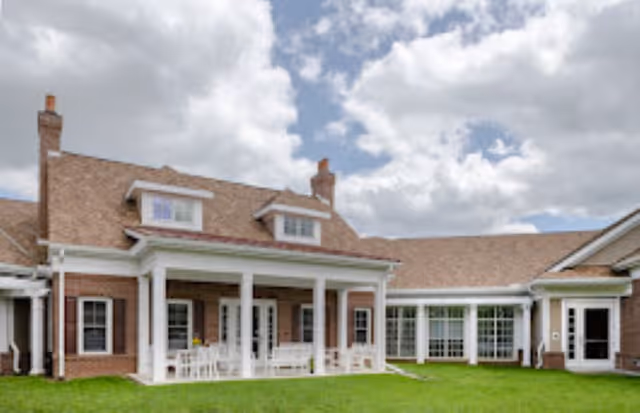 Exterior view of a senior living community building with a brick facade, white columns supporting a covered porch, multiple windows, and a well-maintained green lawn under a partly cloudy sky.