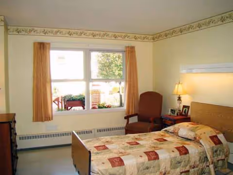 Cozy bedroom inside a senior facility with a bed, chair, dresser, and window view.