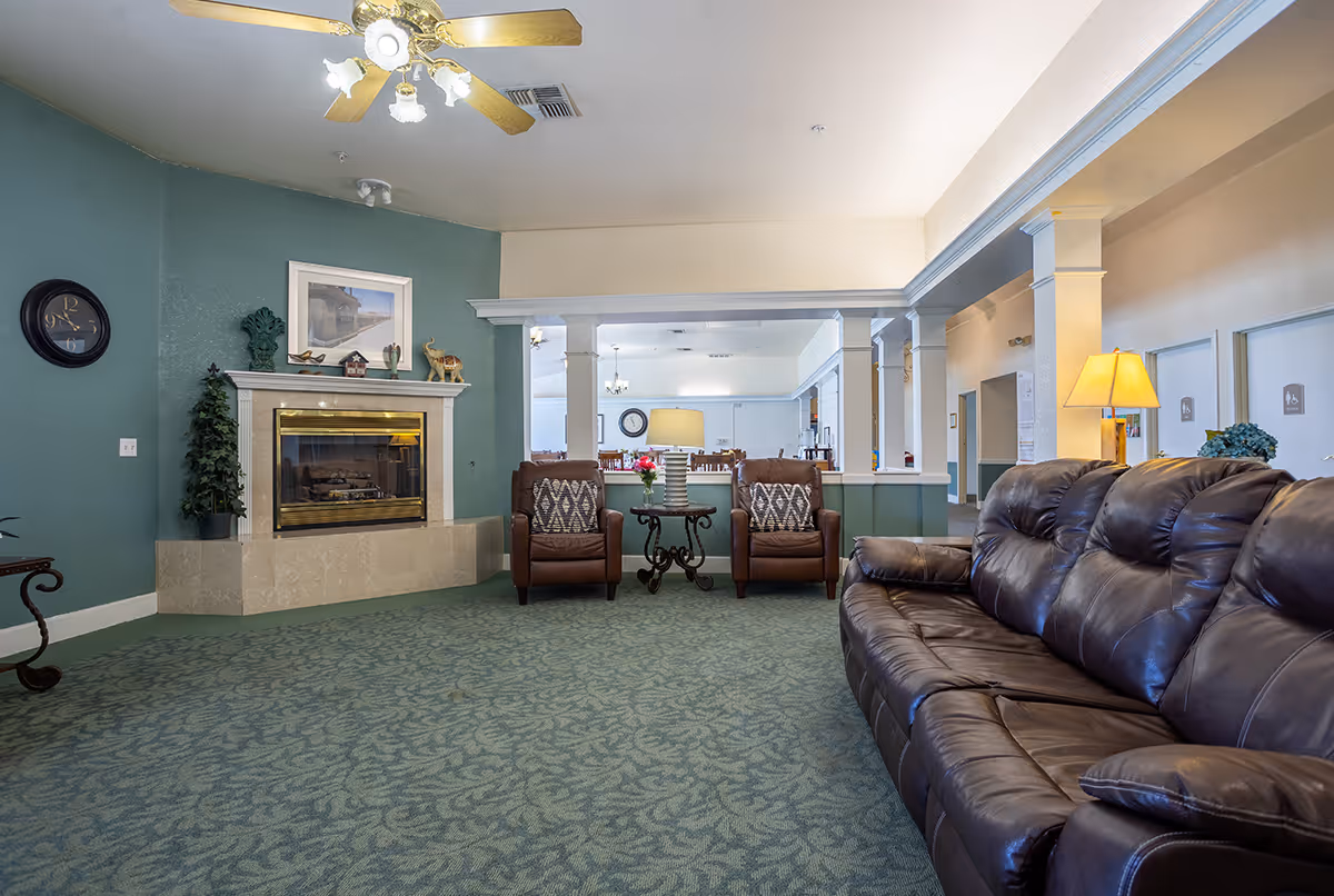 A spacious senior living lounge with a leather sofa, two armchairs, a fireplace, and columns opening to a dining area.