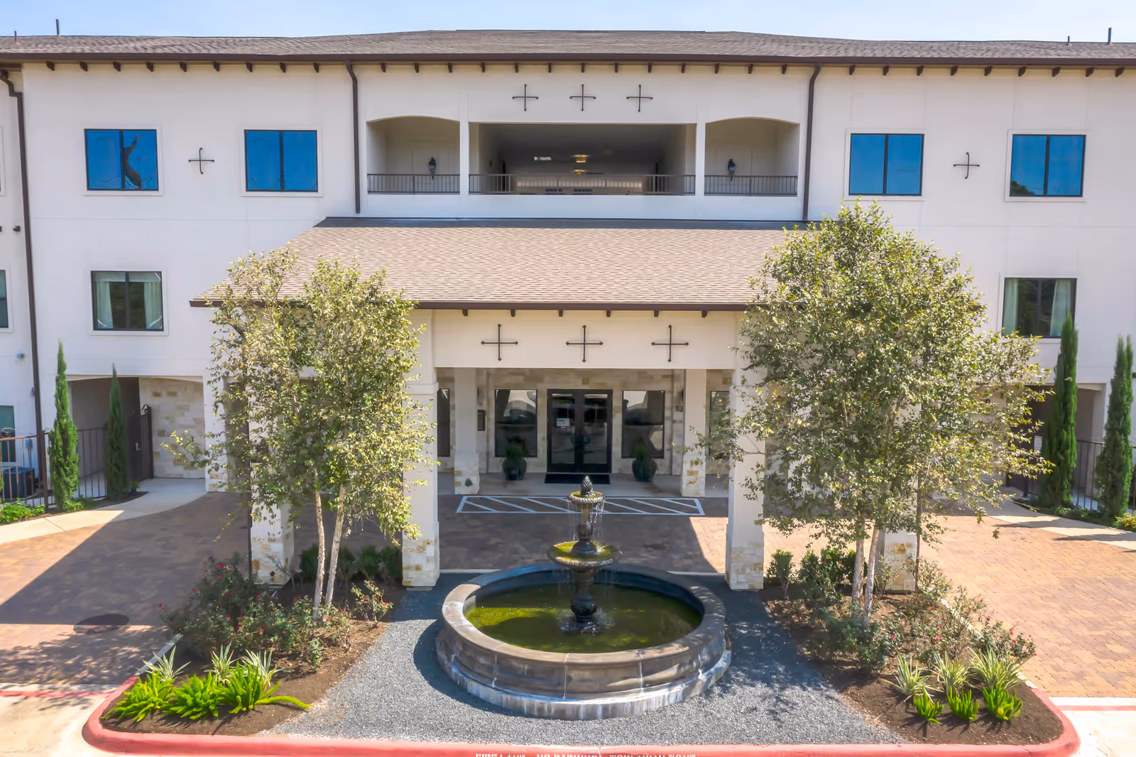 Front exterior view of a senior living facility named Solea Copperfield featuring a covered entrance with stone pillars, a circular water fountain in front, landscaped greenery including trees and shrubs, and a three-story building with multiple windows.