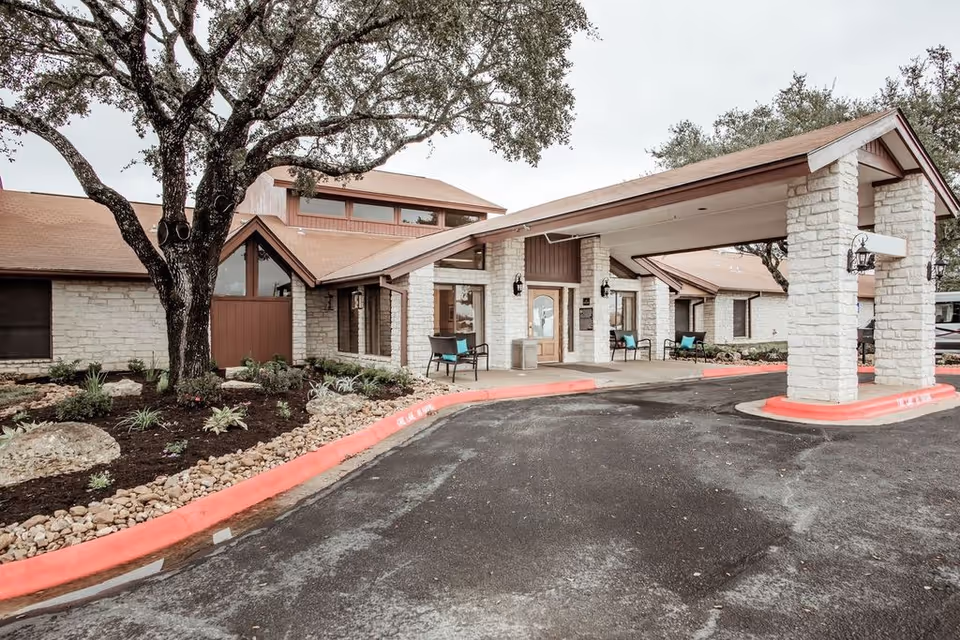 Exterior view of a single-story nursing and rehabilitation center building with stone walls and a covered entrance supported by stone pillars. There are chairs placed near the entrance, a large tree on the left side, and landscaping with rocks and plants along the building. The driveway and curb are visible in the foreground.