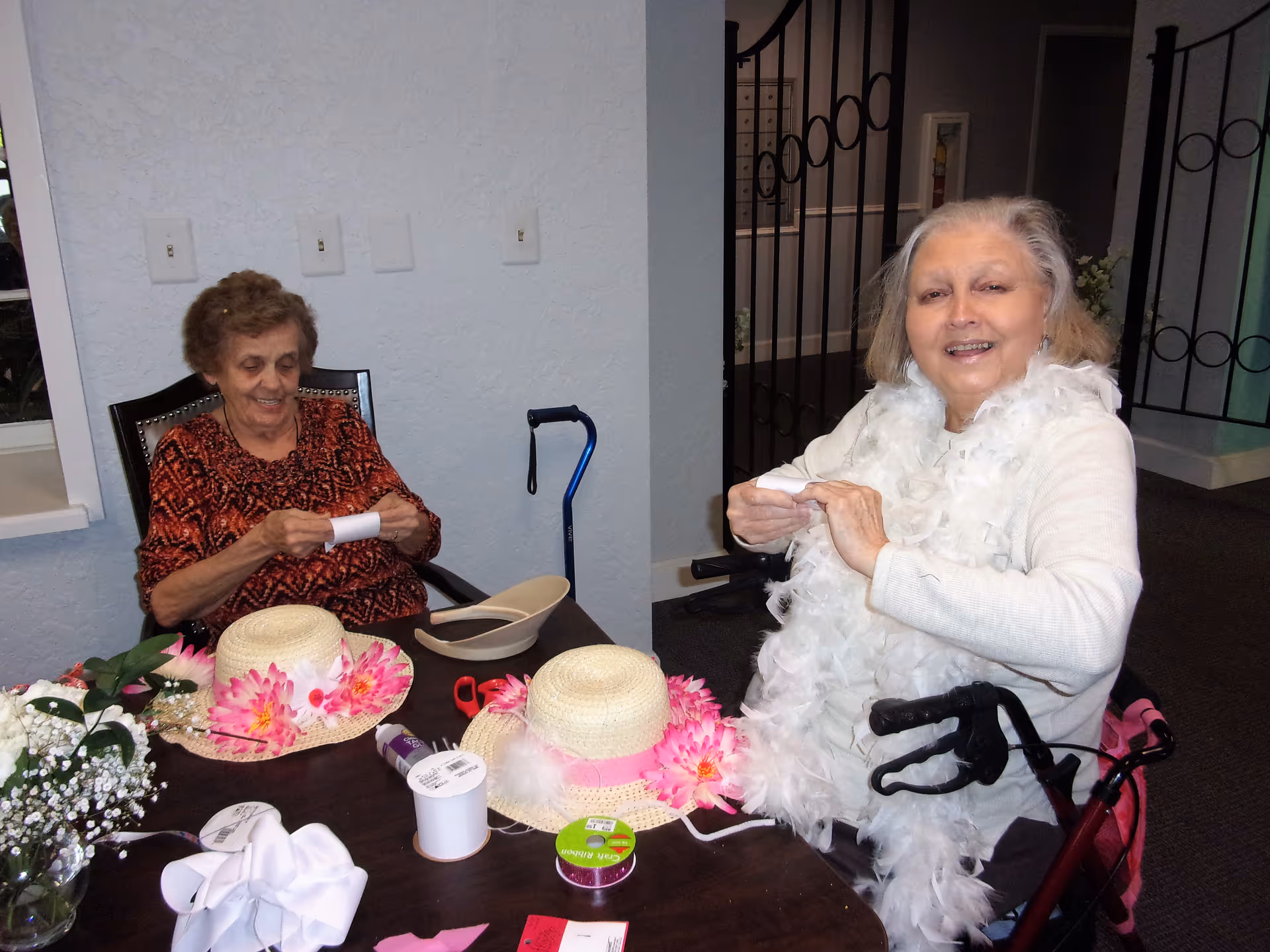 Two elderly women sitting at a table engaged in a craft activity. The table is covered with decorative hats adorned with pink flowers, ribbons, and other craft supplies. One woman is wearing a white feather boa and smiling, while the other is focused on her craft. A blue cane is visible next to the woman on the left.
