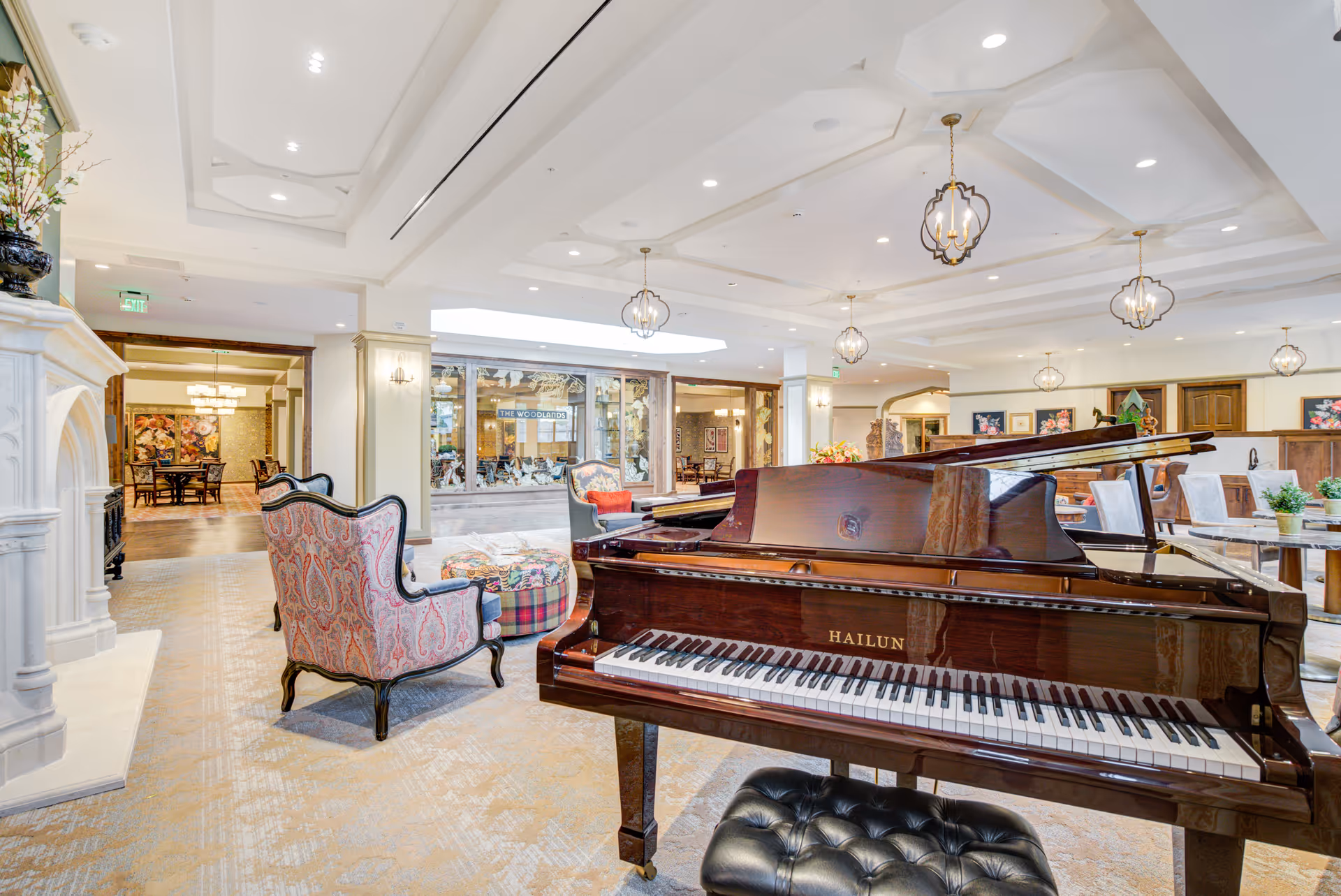 A spacious and elegant living area featuring a polished Hailun grand piano with a black cushioned bench in the foreground. The room includes patterned armchairs, a colorful ottoman, a white fireplace on the left, and several hanging pendant lights. In the background, there is a dining area visible through large windows, and the space is decorated with framed artwork and plants.