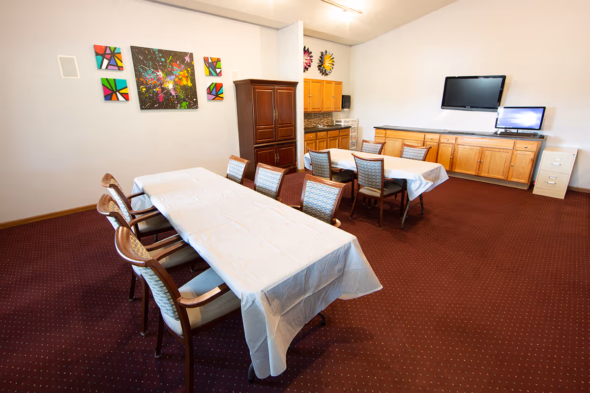 Communal dining/activity room with two long tables covered in white tablecloths, chairs, cabinets, and a wall-mounted TV.