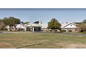 Front view of a single-story senior living facility with a covered entrance, American flag, trees, and a large grassy lawn.