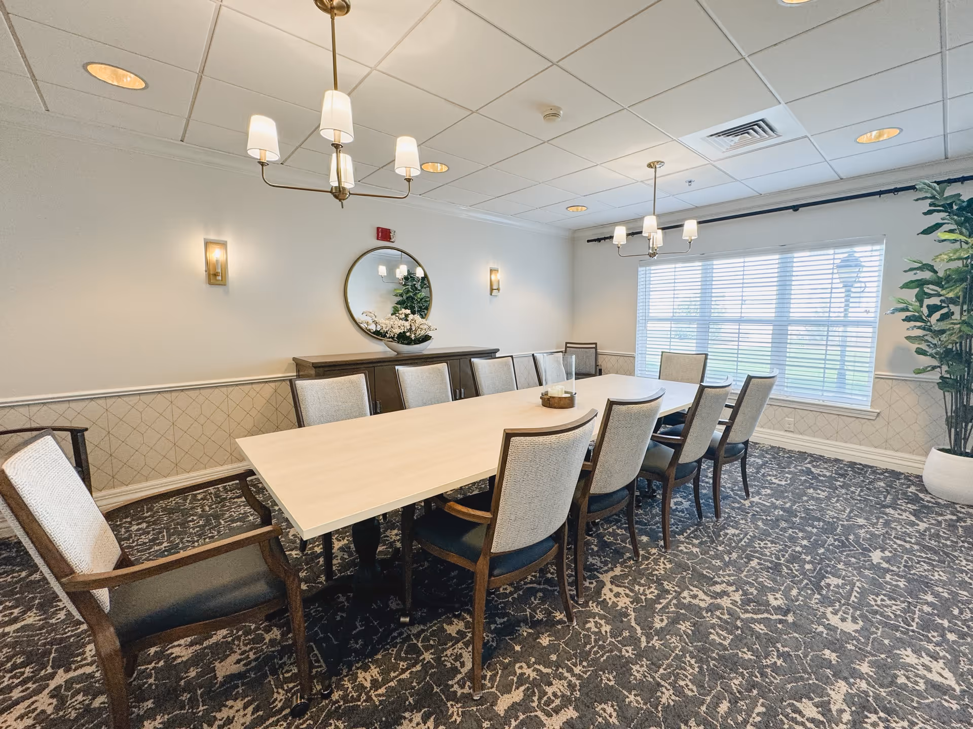 A bright dining room with a long rectangular table surrounded by ten upholstered chairs. The room features patterned carpet, a large window with blinds, two chandeliers, a round mirror on the wall, a sideboard with decorative plants, and wall sconces providing additional lighting.
