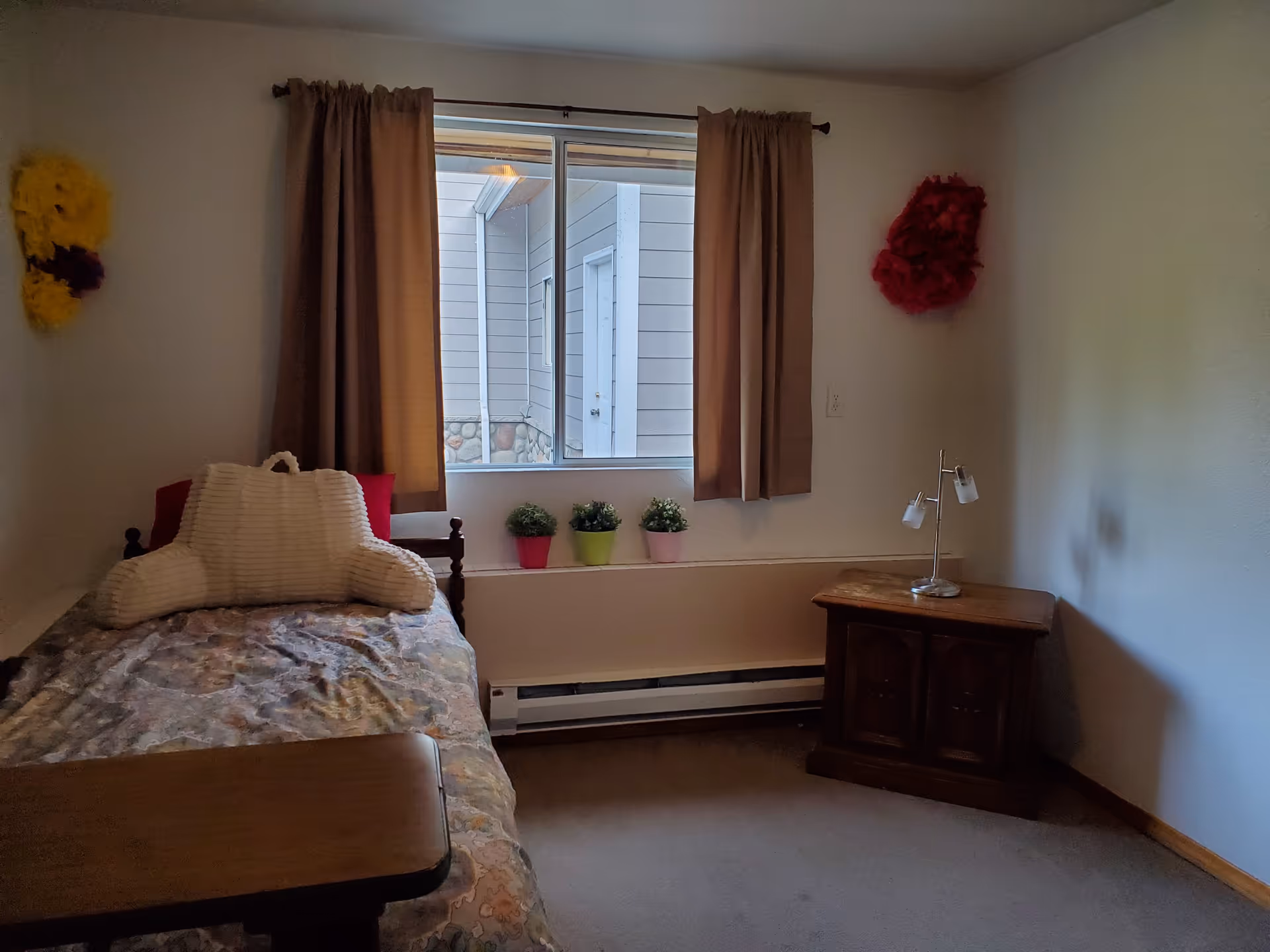 Small furnished single bedroom with a bed, bedside table and lamp, a window with curtains and potted plants on the sill.