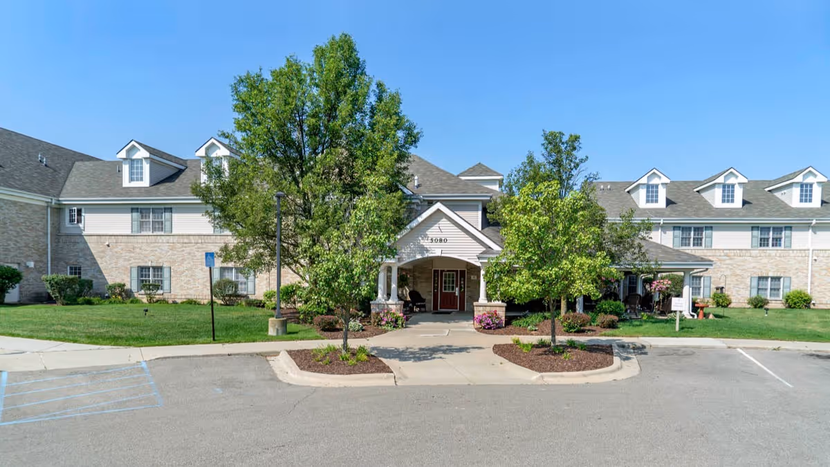Front exterior view of a senior living facility building with a covered entrance, two small trees, and landscaped greenery under a clear blue sky.