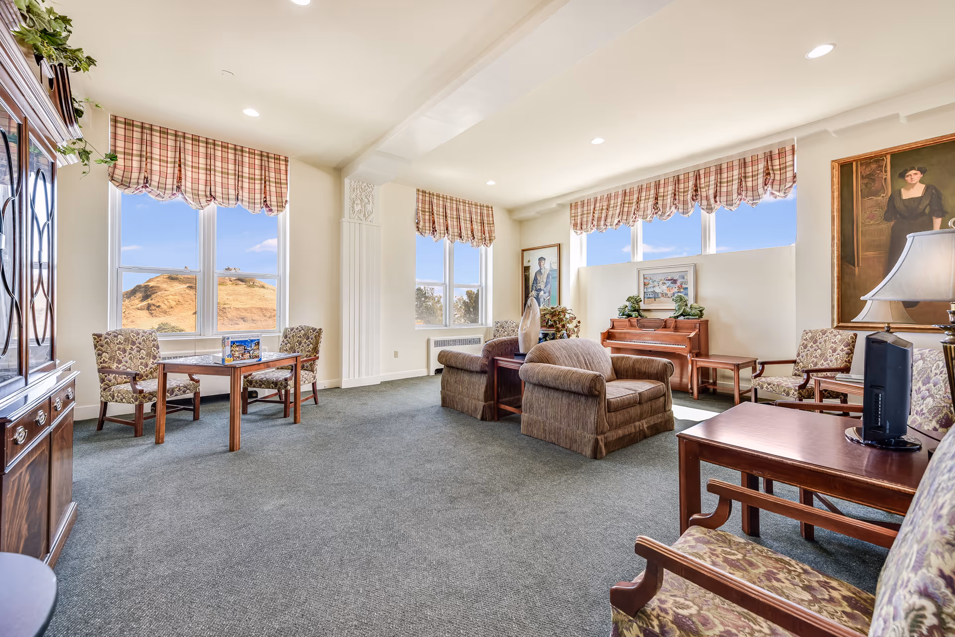 A bright and spacious living room area with large windows featuring plaid valances, allowing natural light to fill the room. The room is furnished with patterned armchairs, a wooden table with a puzzle on it, two cushioned armchairs, a wooden piano, and framed artwork on the walls. The carpet is grayish-blue, and the walls are painted white.