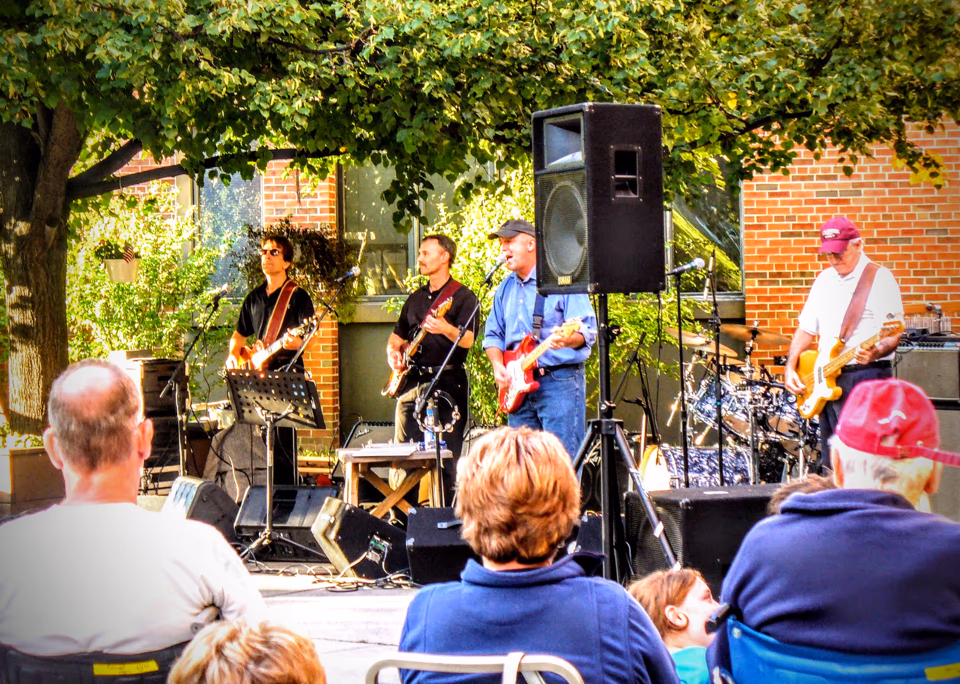 A live band performs outdoors in front of a brick building while seated spectators watch under trees.