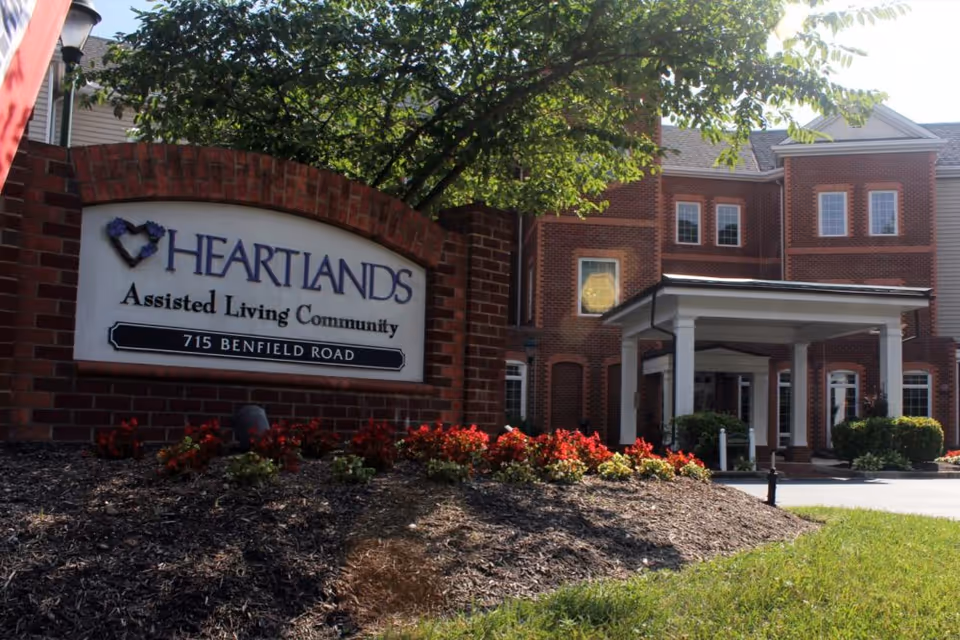 Exterior view of HeartLands Assisted Living Community building with a brick sign displaying the facility name and address 715 Benfield Road, surrounded by landscaping and flowers.